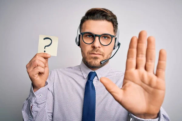 Young call center operator business man with blue eyes holding paper note with question mark with open hand doing stop sign with serious and confident expression, defense gesture