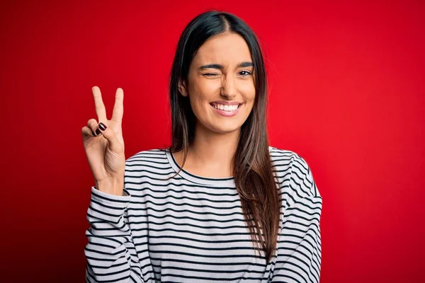 Young beautiful brunette woman wearing casual striped t-shirt over red background smiling with happy face winking at the camera doing victory sign. Number two.