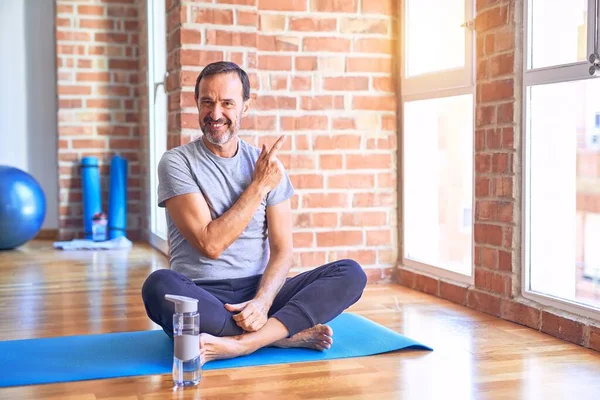 Middle age handsome sportman sitting on mat doing stretching yoga exercise at gym cheerful with a smile on face pointing with hand and finger up to the side with happy and natural expression