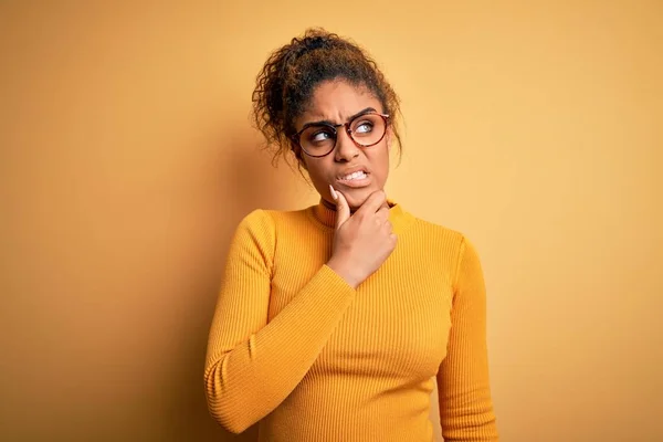 Young beautiful african american girl wearing sweater and glasses over yellow background Thinking worried about a question, concerned and nervous with hand on chin