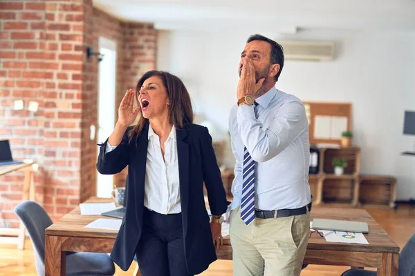 Two middle age business workers standing working together in a meeting at the office shouting and screaming loud to side with hand on mouth. Communication concept.