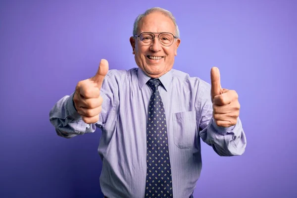 灰白头发的资深商人戴着眼镜 站在紫色孤立的背景上 赞成用手做积极的手势 微笑着竖起大拇指 为成功感到高兴 赢的手势 — 图库照片