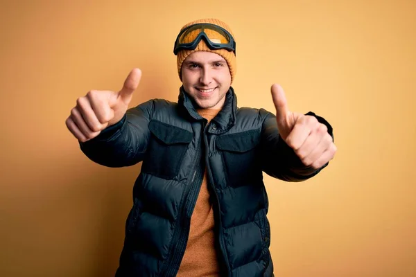 年轻英俊的高加索男子 戴着帽子 外套和滑雪眼镜 在冬天和雪天里 都表示赞成用手做一个积极的手势 微笑着竖起大拇指 为成功感到高兴 赢的手势 — 图库照片
