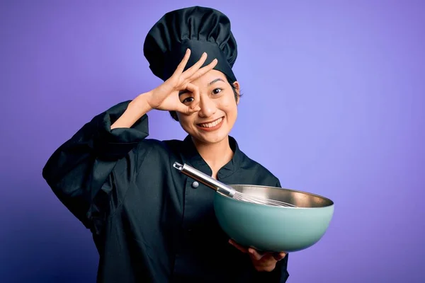 Young beautiful chinese chef woman wearing cooker uniform and hat using bowl and whisk with happy face smiling doing ok sign with hand on eye looking through fingers