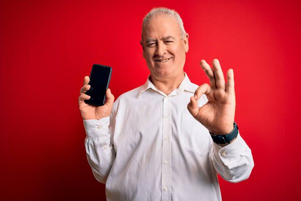 Middle age hoary man holding smartphone showing screen over isolated red background doing ok sign with fingers, excellent symbol