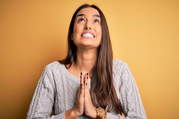 Young beautiful brunette woman wearing white casual sweater over yellow background begging and praying with hands together with hope expression on face very emotional and worried. Begging.
