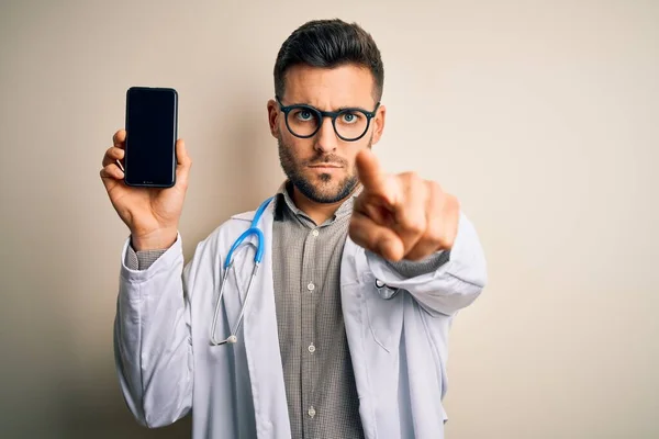 年轻的医生 带着听诊器 在孤立的背景下展示智能手机屏幕 用手指指向相机和你 正面做出积极而自信的手势 — 图库照片