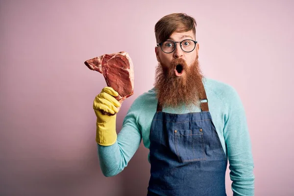 赤毛アイルランドの肉屋男とともに髭を保持生の牛肉ステーキピンクの背景にショックで怖がって驚きの顔 恐怖と興奮で表現 — ストック写真