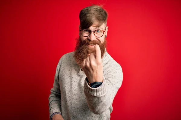 Handsome Irish redhead man with beard wearing casual sweater and glasses over red background Beckoning come here gesture with hand inviting welcoming happy and smiling