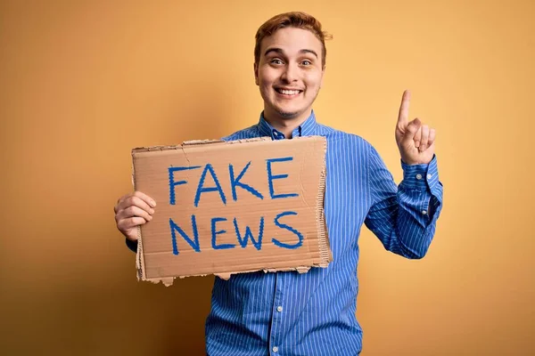 Young handsome redhead man holding banner with fake news message over yellow background surprised with an idea or question pointing finger with happy face, number one