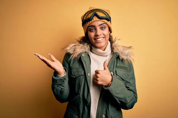 若いアフリカ系アメリカ人のアフロスキーヤーの女の子雪のスポーツウェアとスキーゴーグルを着て手のひらの手を表示し 親指でOkジェスチャーを行うアップ 笑顔と陽気な — ストック写真