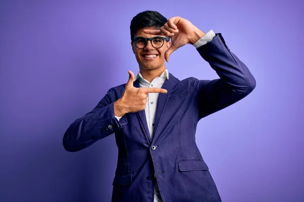 Young handsome business man wearing jacket and glasses over isolated purple background smiling making frame with hands and fingers with happy face. Creativity and photography concept.