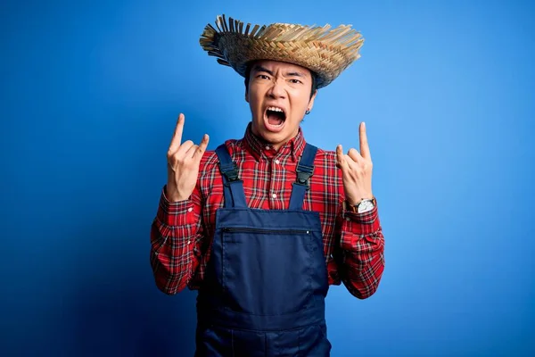 Young handsome chinese farmer man wearing apron and straw hat over blue background shouting with crazy expression doing rock symbol with hands up. Music star. Heavy concept.