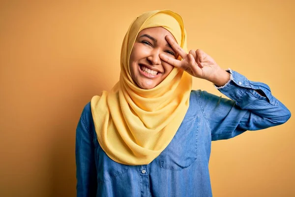 若いです美しいです女性とともに巻き毛身に着けていますアラブ伝統的なヒジャーブ上の黄色の背景を身に着けています顔の上に指で平和のシンボルを行います 笑顔明るい勝利 — ストック写真