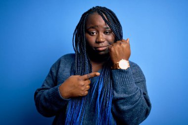 Mavi arka planda günlük süveter giyen Afrikalı Amerikalı artı cüsseli kadın. Aceleyle saati, sabırsızlığı ve rahat bir ifadeyle kameraya bakıyor.