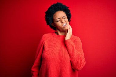 Kıvırcık saçlı, genç Afro-Amerikan kadın diş ağrısı ya da diş ağrısı yüzünden elleriyle dudaklarına dokunan sıradan kazak giyiyor. Dişçi