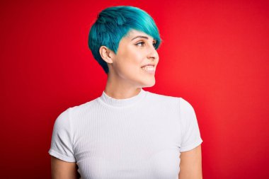 Mavi saçlı, kırmızı arka planda sıradan tişörtler giyen, yüzünde gülümseme, doğal bir ifade olan genç ve güzel bir kadın. Kendinden emin gülen.