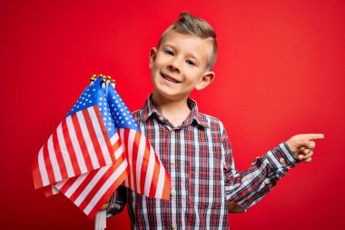 Amerika 'nın küçük vatansever beyaz çocuğu kırmızı arka planda bayrak tutuyor. Elleri ve parmakları kenarda mutlu bir şekilde işaret ediyor.