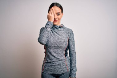 Spor kıyafetleri giyen genç sarışın kadın izole edilmiş arka planda bir gözünü elinle kapatıyor, yüzünde kendine güvenen bir gülümseme ve sürpriz bir duygu var..