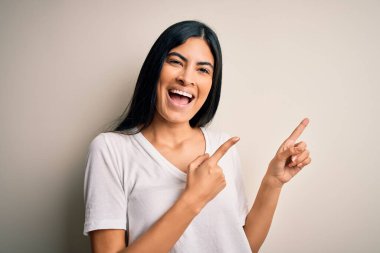 İzole edilmiş arka plan üzerinde gündelik beyaz tişört giyen genç ve güzel İspanyol kadın gülümseyip kameraya bakıyor ve iki eliyle ve parmaklarıyla kenarı işaret ediyor..