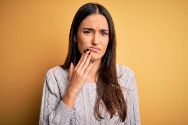 Sarı arka plan üzerine beyaz kazak giyen genç, güzel esmer kadın diş ağrısı ya da diş hastalığından dolayı acı dolu bir ifadeyle ağıza dokunuyor. Dişçi