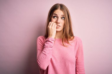 İzole edilmiş pembe arka planda günlük kazak giyen genç, güzel, kızıl saçlı kadın elleri ağız yiyen tırnaklarla gergin ve stresli görünüyor. Endişe sorunu.