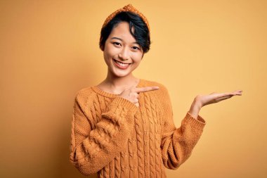 Sarı arka planda dikilen günlük süveter ve diadem giyen genç ve güzel Asyalı kız kameraya hayret ediyor ve gülümsüyordu..