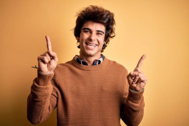 İzole edilmiş sarı arka planda rahat tişört ve süveter giyen genç yakışıklı adam farklı yönleri gösteren kendine güvenle gülümsüyor. Reklam için boşluk kopyala