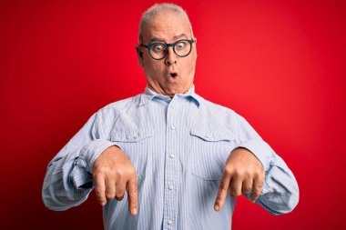 Orta yaşlı, yakışıklı, beyaz gömlekli, kırmızı arka planda gözlüklü, beyaz bir adam. Parmaklarını göstererek reklamını yapıyor, şaşırmış yüzünü ve ağzını açıyor.