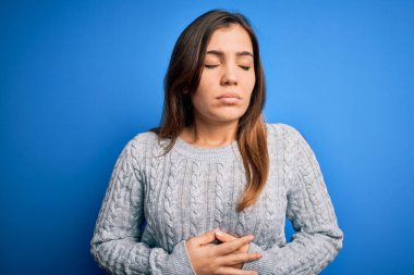 Güzel, genç bir kadın günlük yün kazak giyiyor. Mavi yalıtılmış arka plan üzerinde duruyor. Midenin üstünde elleri var. Ache kavramı.