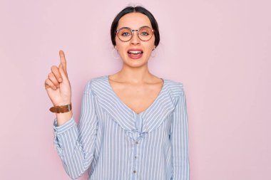 Pembe arka planda rahat çizgili gömlek ve gözlük takan genç ve güzel bir kadın bir numaralı parmağını gösterirken kendine güvenen ve mutlu bir şekilde gülümsüyor..