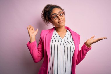 Güzel Afro-Amerikalı iş kadını pembe arka planda ceket ve gözlük takıyor ve elleri ve kolları havada, kafası karışmış bir ifade takınıyor. Şüphe kavramı.