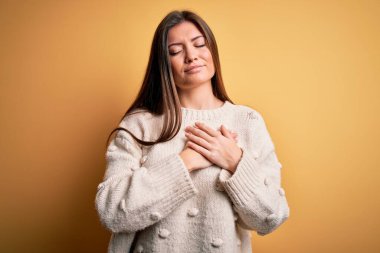 Mavi gözlü genç bir kadın sarı arka planda sıradan bir süveter giyiyor. Elleri göğsünde, gözleri kapalı ve yüzünde minnet dolu bir jest ile gülümsüyor. Sağlık kavramı.