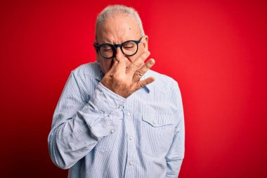 Orta yaşlı, beyaz, sıradan çizgili gömlek ve kırmızı arka planda gözlük takan yakışıklı adam pis ve iğrenç bir koku alıyor. Nefesini burnunda tutuyor. Kötü kokuyor.