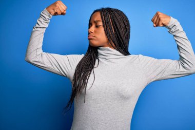 Genç Afro-Amerikalı kadın, mavi izole arka plan üzerine balıkçı yaka kazak giyip gururla gülümseyen kol kaslarını gösteriyor. Fitness konsepti.