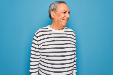 Kıdemli yakışıklı, gri saçlı, çizgili süveter giyen, soyutlanmış mavi arka planda, yüzünde gülümseme ve doğal bir ifadeyle bir yana bakan adam. Kendinden emin gülen.