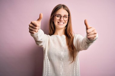 Genç, güzel, kızıl saçlı bir kadın pembe arka plan üzerine günlük süveter ve gözlük takarak olumlu bir jest yapmayı onaylıyor. Elleriyle gülümsüyor ve başarı için mutlu. Kazanan jest.