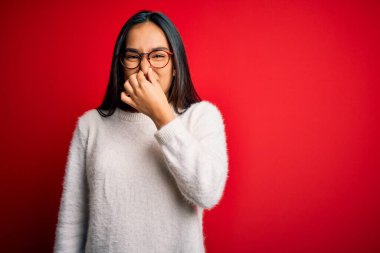 Genç ve güzel Asyalı bir kadın kırmızı arka planda günlük süveter ve gözlük takıyor. Kokuşmuş ve iğrenç bir koku alıyor. Dayanılmaz bir koku. Kötü kokuyor.