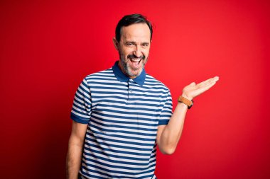 Orta yaşlı, beyaz, sıradan çizgili polo giyen, izole edilmiş kırmızı arka planda duran neşeli bir sunum yapan ve avuç içi kameraya bakan bir adam..