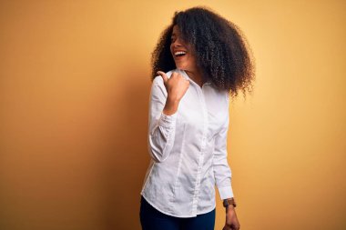 Afro saçlı, sarı arka planda gülümseyen, gülümseyen ve baş parmağıyla yüzünü gösteren güzel Afro-Amerikan zarif bir kadın..