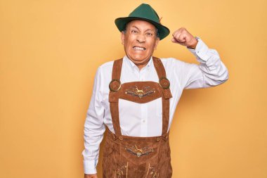 Kıdemli gri saçlı adam, sarı arka plan üzerine geleneksel Alman Ekim Festivali kıyafeti giymiş kızgın ve öfkeyle bağırırken yumruğunu kaldırmış öfkeli. Öfke ve agresif kavram.