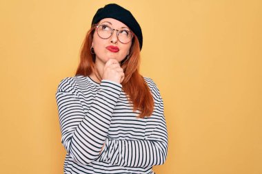 Sarı arka planda çizgili tişört ve Fransız beresi giyen güzel kızıl saçlı bir kadın. Elleri çenesinde soru sorarken, düşünceli bir ifade takınıyor. Düşünceli bir yüzle gülümsüyor. Şüphe kavramı.