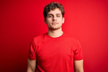 Sarışın, kıvırcık saçlı, kırmızı arka planda sıradan tişörtler giyen, yüzünde ciddi bir ifadeyle rahatlamış genç bir adam. Basit ve doğal bir şekilde kameraya bakıyorsun..