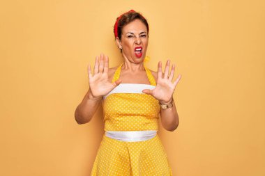 Orta yaşlı bir kadın, 50 'li yaşlarda, sarı arka planda eski tarz bir elbise giyiyor. El hareketlerini, kızgın ve hüsran dolu ifadelerini durduruyor.