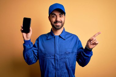 Sakallı, mavi üniformalı ve şapkalı tamirci elinde akıllı telefonuyla ekranı çok mutlu bir şekilde işaret ediyor.