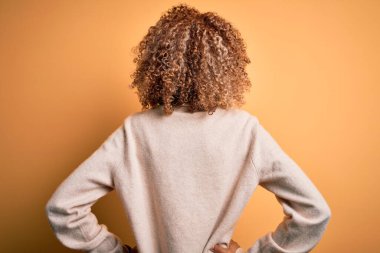 Güzel, Afro-Amerikan kadın balıkçı yaka kazak giyiyor sarı arka planda geriye bakıyor kollarını vücuduna dikiyor.