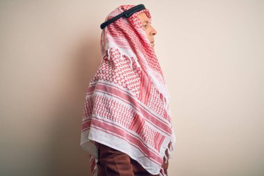 Kıdemli yakışıklı Arap adam, keffiyeh giyip izole edilmiş beyaz arka planda duruyor. Yan tarafa bakıyor, rahat bir görünüm sergiliyor, kendine güvenen bir gülümsemeyle doğal bir yüz ifadesi sergiliyor..