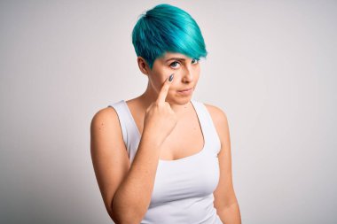 Mavi saçlı, beyaz arka planda sıradan tişörtler giyen genç ve güzel bir kadın jestini ve şüpheli ifadeni izliyor.