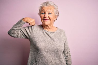 Üst düzey güzel bir kadın, günlük tişört giyip pembe arka planda dikiliyor. Güçlü biri kol kası gösteriyor, kendinden emin ve gururlu.