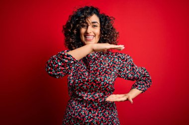 Genç, güzel, kıvırcık, çiçekli bir elbise giyen, kırmızı arka planda duran, elleri büyük ve büyük bir işaret gösteren, ölçü sembolü olan, kıvırcık bir kadın. Kameraya bakıp gülümsüyor. Ölçüm konsepti.